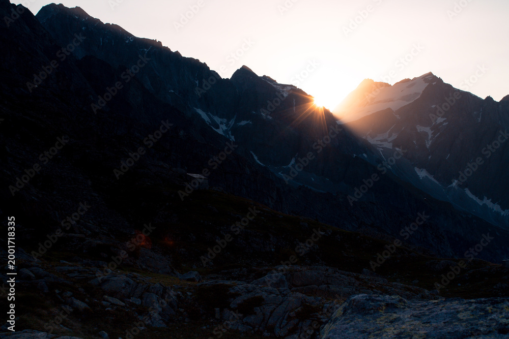 Obraz premium Vibrant sunstar bursting over dark alpine mountain ridge with colorful lens flare, turquoise sky and rocky foreground, Alps Italy