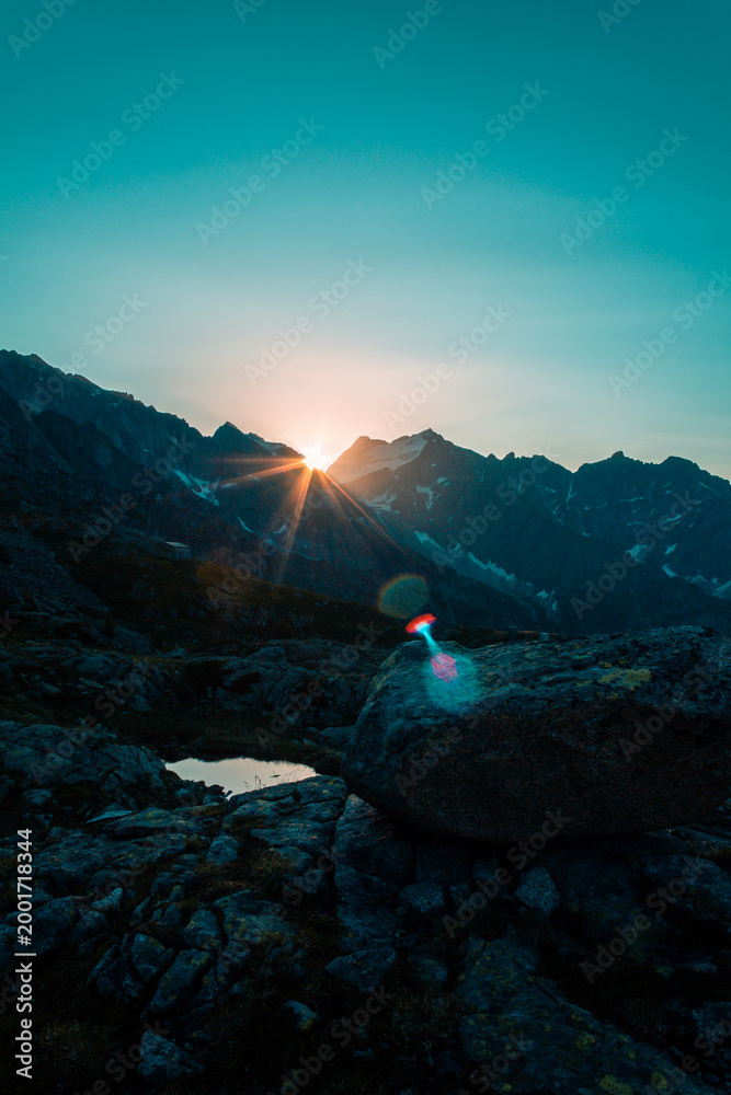 Obraz premium Intense sunburst with dramatic lens flare over dark alpine mountain silhouette, deep blue sky and rocky terrain at sunrise, Italian Alps