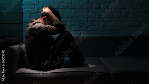Young man sitting on a dark sofa with his head in his hands, lonely and anxious, struggling with depression and emotional distress in a moody, low key interior
