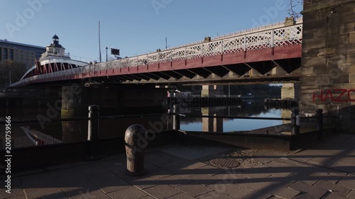 Ground-level pan of the historic Swing Bridge in Newcastle at dawn, highlighting the industrial iron structure and stone pillars under the low early morning sun.
