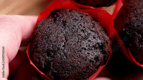 Close-up of a person hand picking up a tempting chocolate muffin in a red wrapper. Macro texture of porous cocoa sponge cake on wooden table