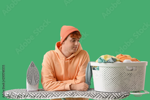 Tired young man with electric iron and full laundry basket leaning on ironing...