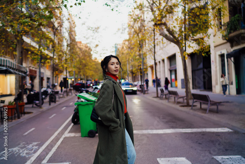 Fotografie Multiracial woman in olive coat looking back while crossing street, young female traveler in autumn city setting with soft bokeh background, authentic representation of urban exploration