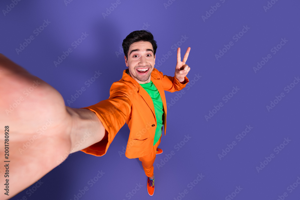 Fototapeta premium Happy young man in orange suit taking a high-angle selfie with peace sign on purple background, colorful fashion portrait and playful studio mood