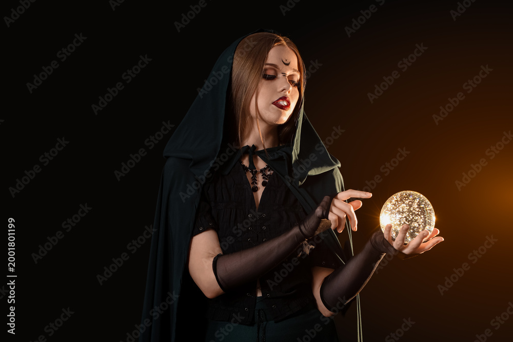 Fototapeta premium Beautiful young witch with crystal ball on dark background