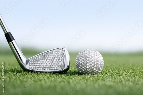 A silver golf club head covered in small sparkling diamonds positioned next to a white dimpled golf ball on a lush green grass golf course fairway