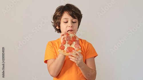 A boy in an orange shirt holds a large pizza slice near his mouth, captured from the front with a clean background. Convenience food, kids appetite, and family dining reflect modern lifestyle choices.