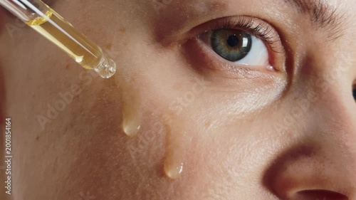 Woman applying liquid skincare serum with a pipette to her face, showing large droplets running down her cheek, emphasizing hydration, healthy skin, and anti aging cosmetic treatment
