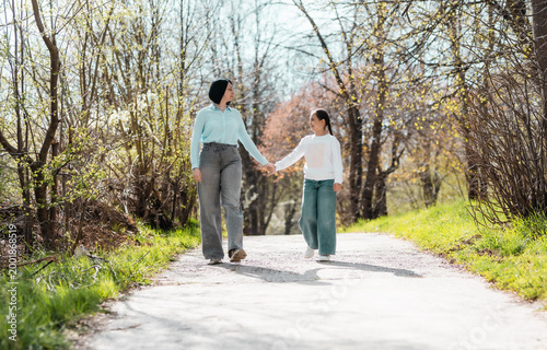 Happy mother and little daughter holding hands and walking in the park, looking at each other with love and affection