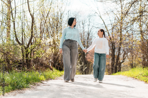 Happy mother and little daughter holding hands and walking in the park, looking at each other with love and affection