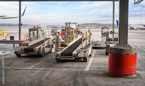 Belt loaders and ground support equipment stand ready on the airport tarmac with an airplane visible in the background.