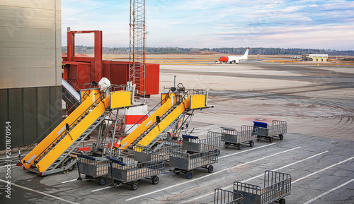 Ground support equipment including yellow passenger stairs and baggage carts sit on an airport apron with an airplane taxiing in the background under a soft sunset sky.