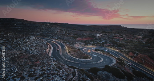Malta: Aerial view of cars driving on a winding road on the coast of Malta at bright sunset, creating a scenic landscape with the Mediterranean Sea in the background. Drone flight