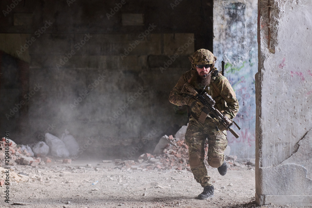 Fototapeta premium Armed Soldier In Camouflage Tactical Gear Running Through Urban Ruins With Rifle Ready
