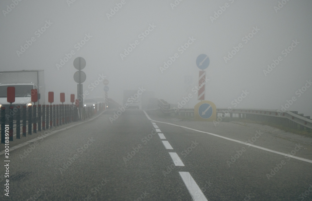 Fototapeta premium Foggy road with cars and warning signs in low visibility.