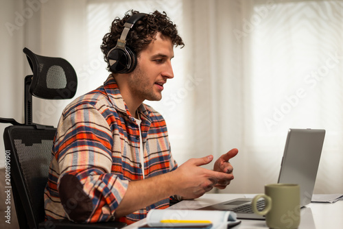 Young man using laptop with headset, participating in online class and virtual meeting. Remote work and e-learning concept in a home office environment.