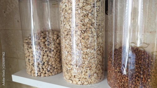 Transparent jars holding black-eyed peas, rolled oats, and buckwheat groats are neatly arranged on a white shelf, showcasing efficient kitchen pantry organization and healthy food storage