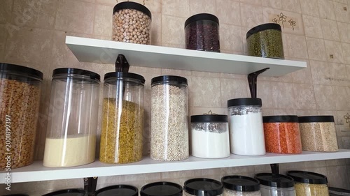Kitchen pantry shelves filled with various legumes, grains, and flour stored in transparent containers, presenting an organized and practical kitchen storage solution