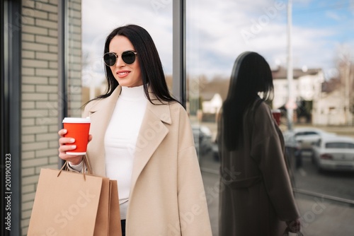 Foto Woman in beige coat holding coffee and shopping bags with sunglasses by shop win