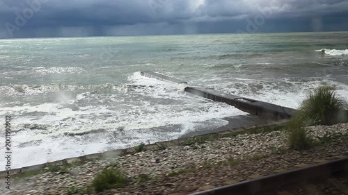 Turbulent sea with powerful waves crashing onto the shore next to a railway line, filmed from the window of a moving train under a dramatic, dark, and cloudy sky on a stormy day