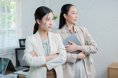  Two professional businesswomen are working on laptop computers sitting at desks in the office. Colleagues are happy and discussing the digital strategy of the Graph