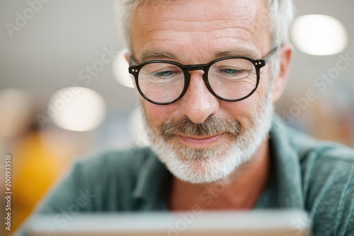 Focused senior man with grey hair and white beard wearing glasses looking at tablet screen indoors