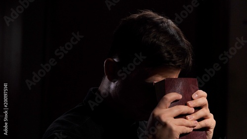 Foto Man resting his forehead on a red book held in both hands in dim light, a solemn