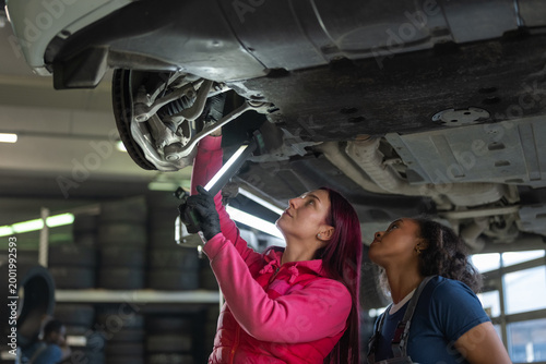 Two women car mechanics using a torch and a digital tablet while checking the undercarriage of a vehicle in a busy repair shop.