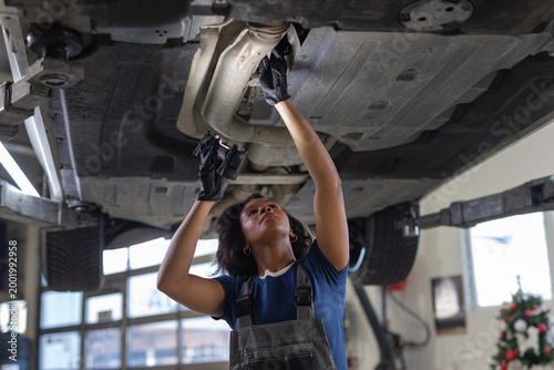 Young African American female mechanic using a flashlight to inspect the underside of a car raised on a lift in an auto repair shop.