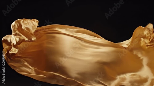 A close-up shot showcases the flowing, golden texture of draped fabric against a dark background, highlighting its luxurious and dynamic form.