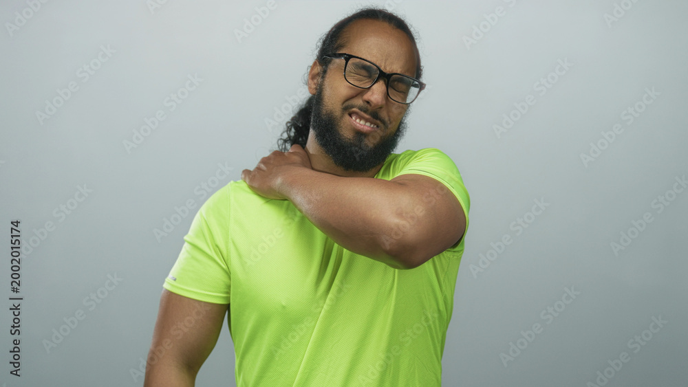 Fototapeta premium Man clutching shoulder and wincing while wearing neon green shirt in gray studio after intense workout session; discomfort.