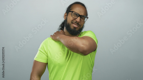 Man clutching shoulder and wincing while wearing neon green shirt in gray studio after intense workout session; discomfort.