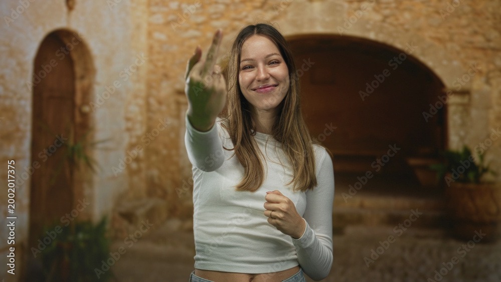 Fototapeta premium Woman shows middle finger toward camera on an old town street, wearing white top and jeans; rebellion defiance.
