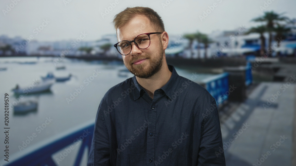 Fototapeta premium Man smiling with glasses and visible face on a seaside street promenade, hands relaxed; serenity confidence.