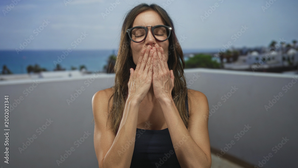 Fototapeta premium Woman blows a kiss through black glasses with both hands on a seaside balcony overlooking the ocean; affection longing.
