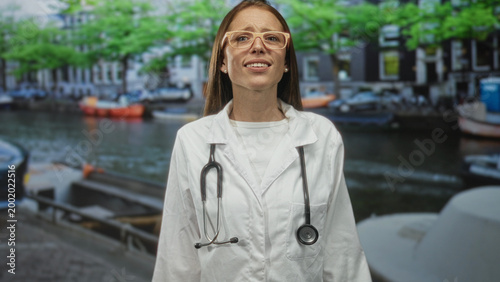 Принти на полотні Young woman doctor in white coat with stethoscope raising hands on canal street in amsterdam by moored boats and waterway; medical care concern