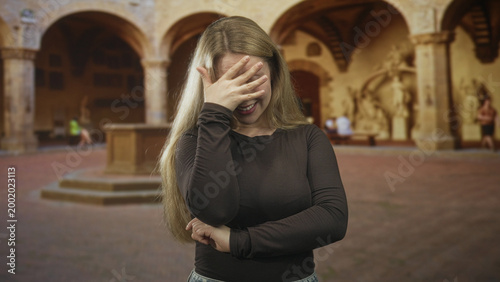 Εκτύπωση καμβά Woman blonde covers face with hand and presses forehead, standing in a historic courtyard inside a building with arches and statues; embarrassment introspection