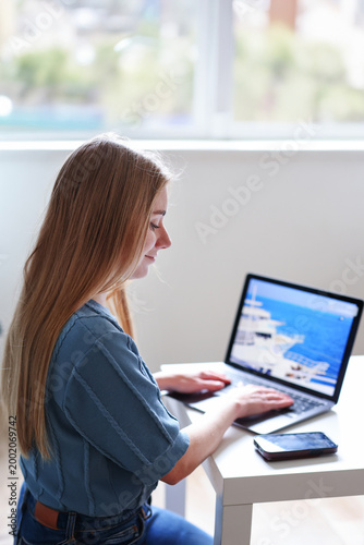 Female freelancer using laptop and smartphone at home planning vacation online. Remote work and travel concept.