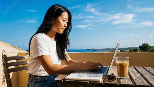 Asian woman working on laptop at outdoor cafe table by sea. Freelancer typing on computer during sunny day. Remote work lifestyle and digital nomad concept.