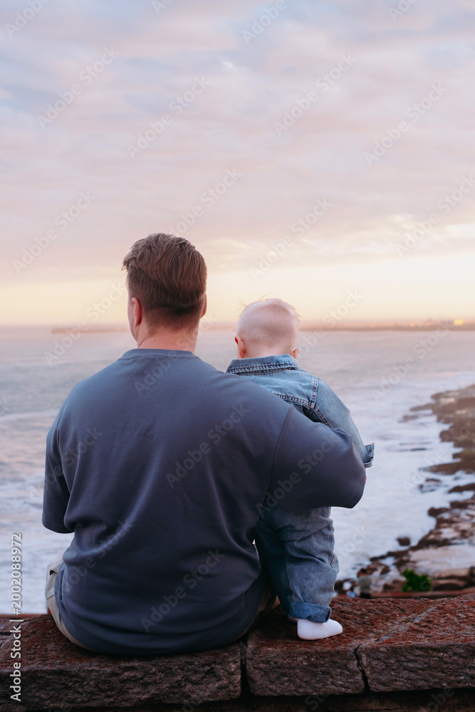 Fototapeta premium Father holding baby and looking at the sea. Calm emotional moment of parenting, family bonding, and peaceful lifestyle by the water.