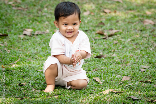 A cheerful child in white clothing sits smiling on the grass in an outdoor setting