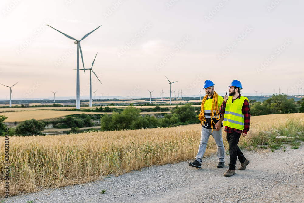 Obraz premium Engineers in hard hats and safety vests walking on a dirt road, discussing operations at a wind farm generating clean energy. Renewable energy and industrial workforce concept