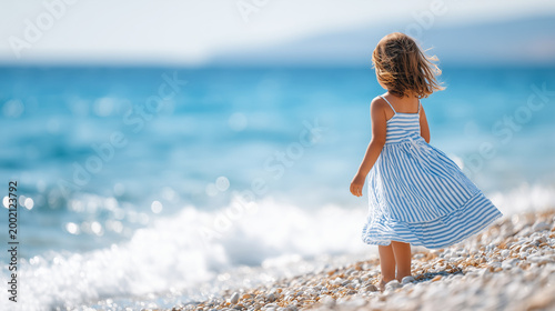Little girl in a blue striped dress standing on a sunny pebble beach by the sea with waves and wind in her hair.