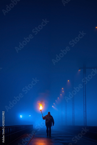 Silhouette of a lone person walking on a foggy night street holding a burning torch with blue mist and street lights glowing in the distance