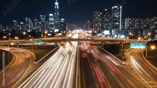 A long exposure photograph captures the vibrant flow of traffic on a highway at night, with a stunning cityscape illuminated in the background.