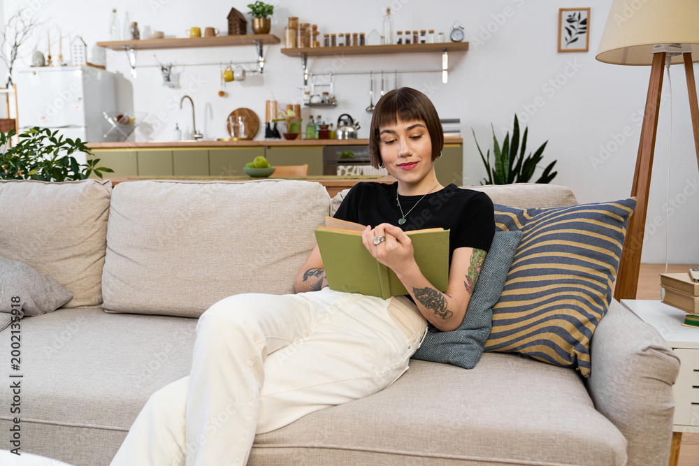 Obraz premium Young woman enjoys a cozy afternoon reading a book in her modern living room