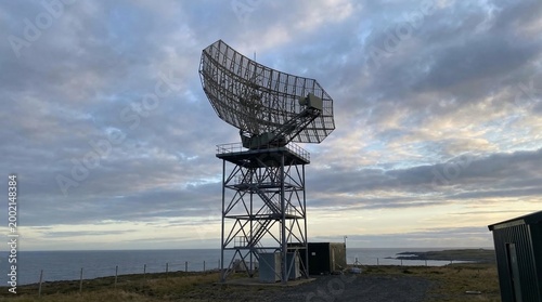 Large radar antenna on a metal tower overlooking the ocean under a cloudy sky