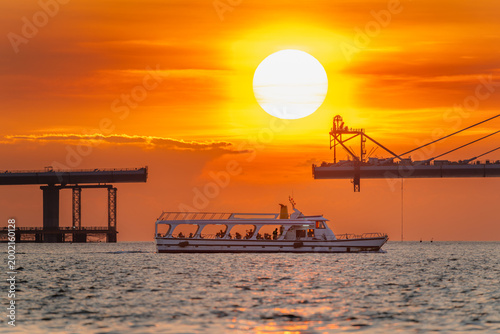 A scenic view of the Danjiang Bridge construction site in New Taipei City, Taiwan, during a vibrant summer sunset with a passenger ferry boat passing by.