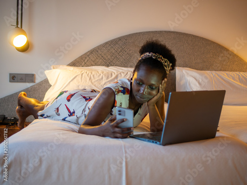Obraz na plátně Smiling African woman typing on her smartphone while using her laptop lying prone on a cozy bed