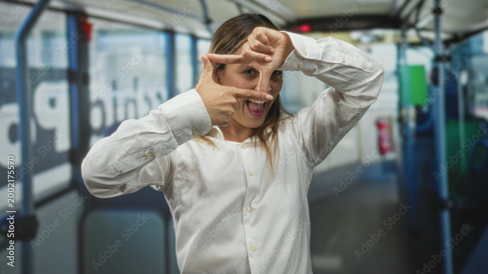 Obraz premium Woman framing a rectangle with fingers inside a bus interior on a street, wearing a white shirt and focusing on hands; playful focus.